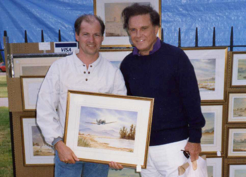 Bill Holkham standing at his large outdoor art stall displaying framed watercolor paintings
